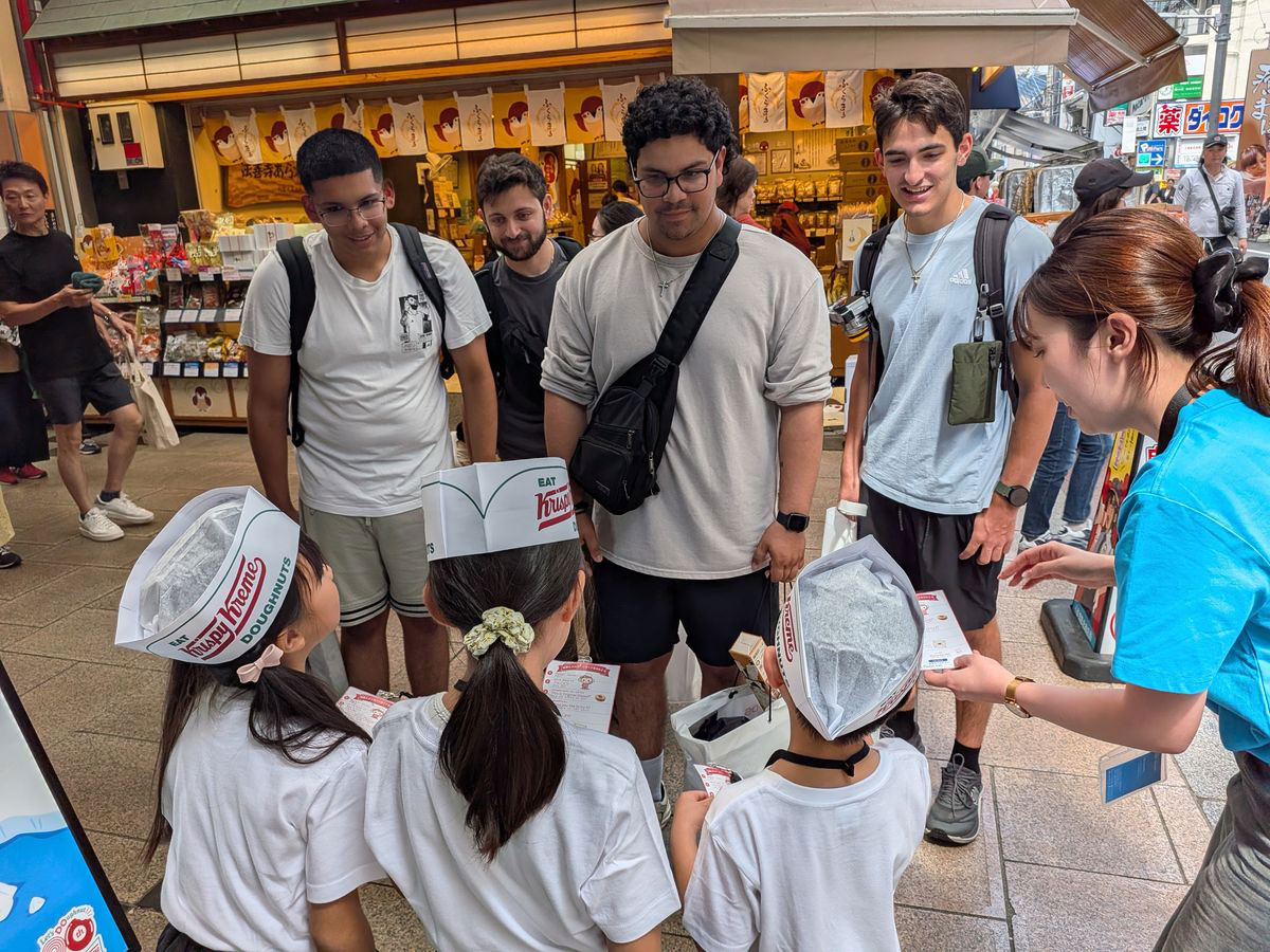 商店街の店頭でクリスピー・クリーム・ドーナツを外国人観光客に英語でおすすめする子どもたち
